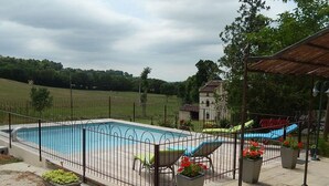 Une piscine rectangulaire entourée d’une clôture métallique, avec une terrasse dallée où se trouvent des chaises longues et des plantes en pot.