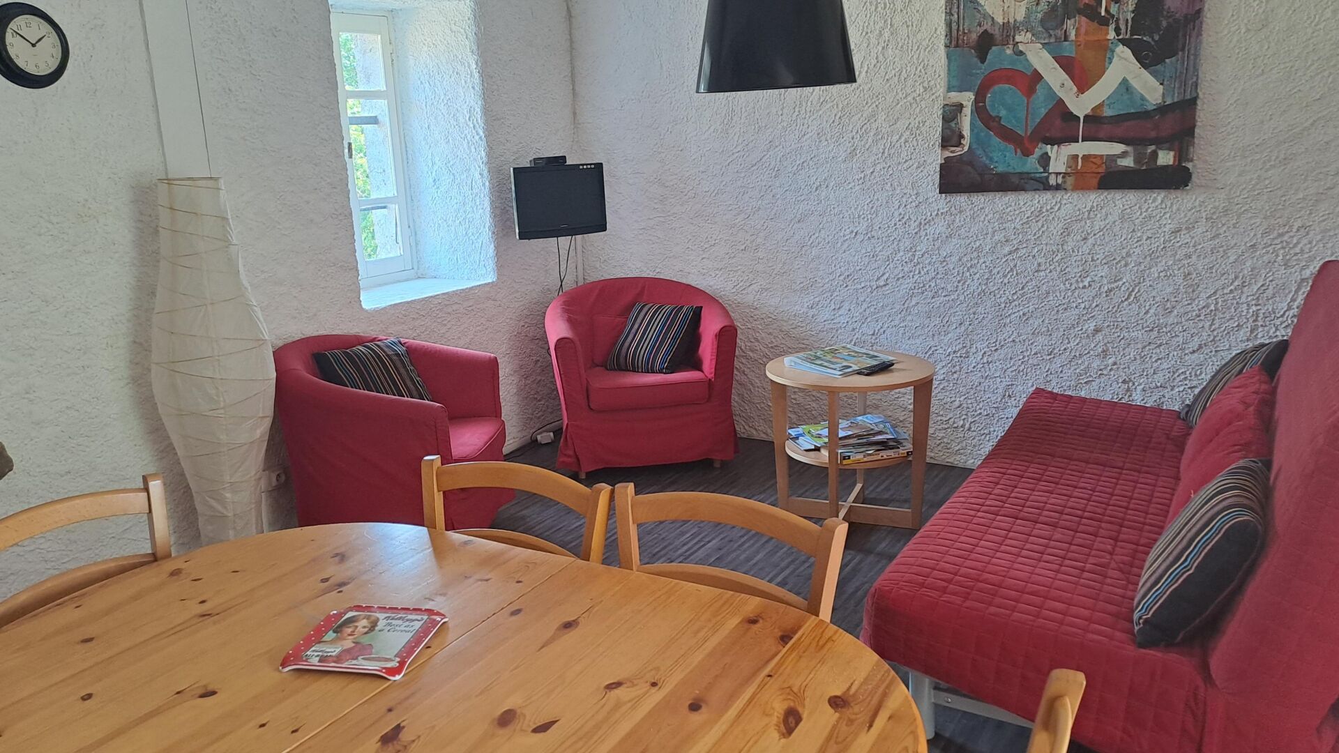 Un salon chaleureux avec une table en bois, des fauteuils rouges, un canapé rouge, une petite table d’appoint et un téléviseur à écran plat fixé au mur.