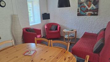 Un salon chaleureux avec une table en bois, des fauteuils rouges, un canapé rouge, une petite table d’appoint et un téléviseur à écran plat fixé au mur.