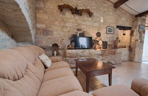 Ett rustikt vardagsrum med väggar av natursten, en beige soffa, ett soffbord i trä och en tv som står på en hylla av sten.