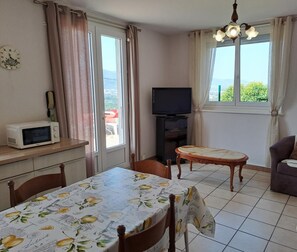 A small, well-lit room with a dining table, chairs, a sofa, a television, and a window with curtains.