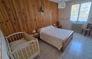 Un dormitorio con pared de paneles de madera, una cama con colcha floral, una mesita de noche con lámpara, una silla de madera y un radiador.