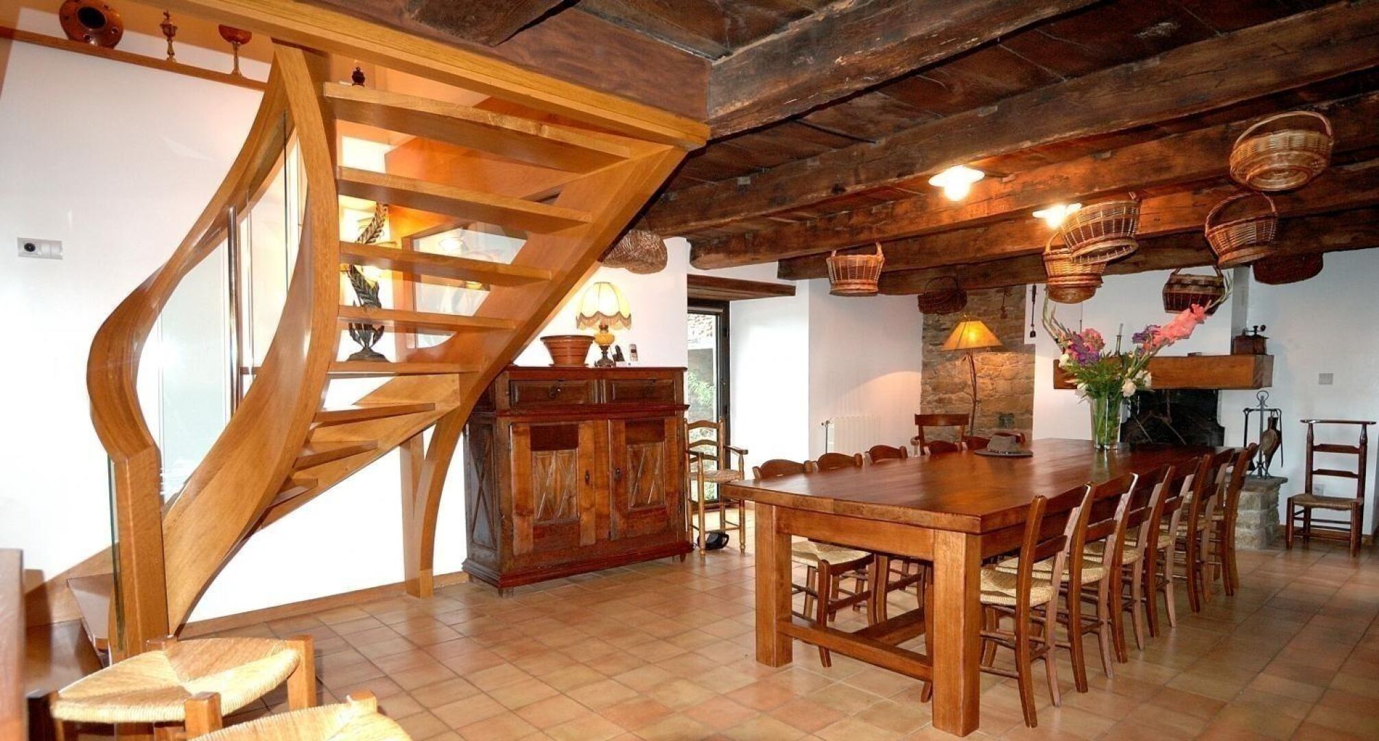 A rustic dining area with a wooden table and chairs, a spiral staircase, and hanging baskets.