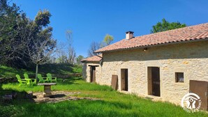 A stone building with a tiled roof, a stone bench, and green chairs in a grassy area with trees in the background.