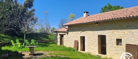 A stone building with a tiled roof, a stone bench, and green chairs in a grassy area.