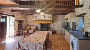A rustic kitchen with a washing machine, wooden chairs, and a dining table with a patterned tablecloth.