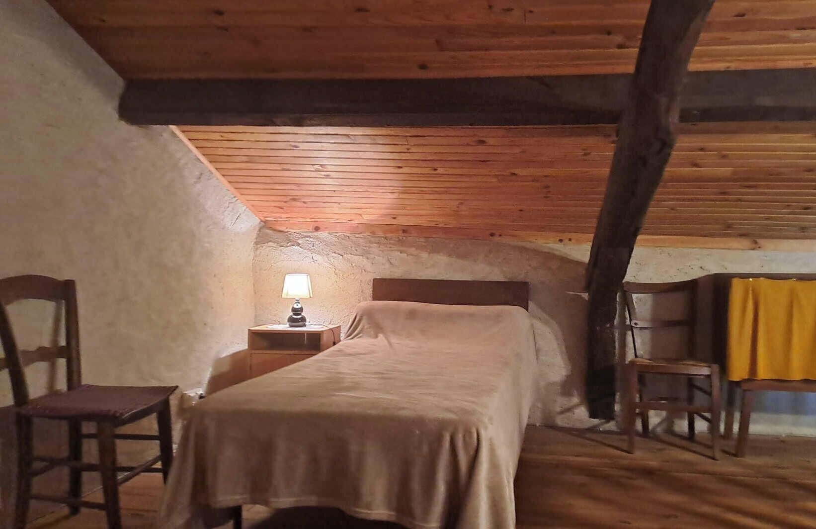A cozy attic bedroom with a wooden ceiling, a single bed, a nightstand with a lamp, and a chair.