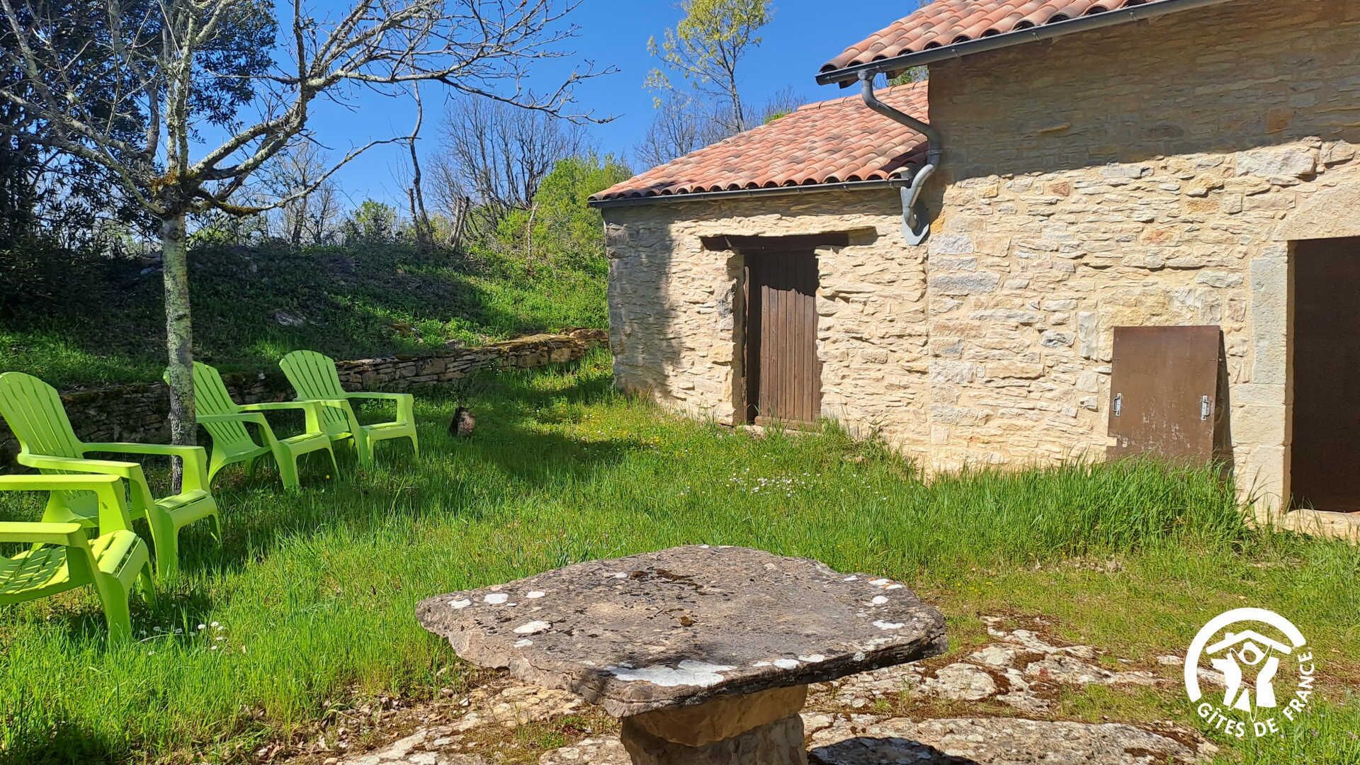 Un edificio in pietra con tetto in tegole, una porta in legno e una panchina in pietra in un'area erbosa con sedie verdi.