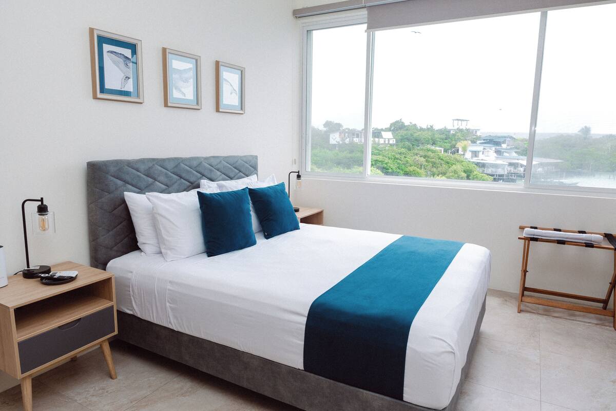 A modern bedroom with a large bed, a bedside table, a nightstand, and a window with a view of greenery and buildings.