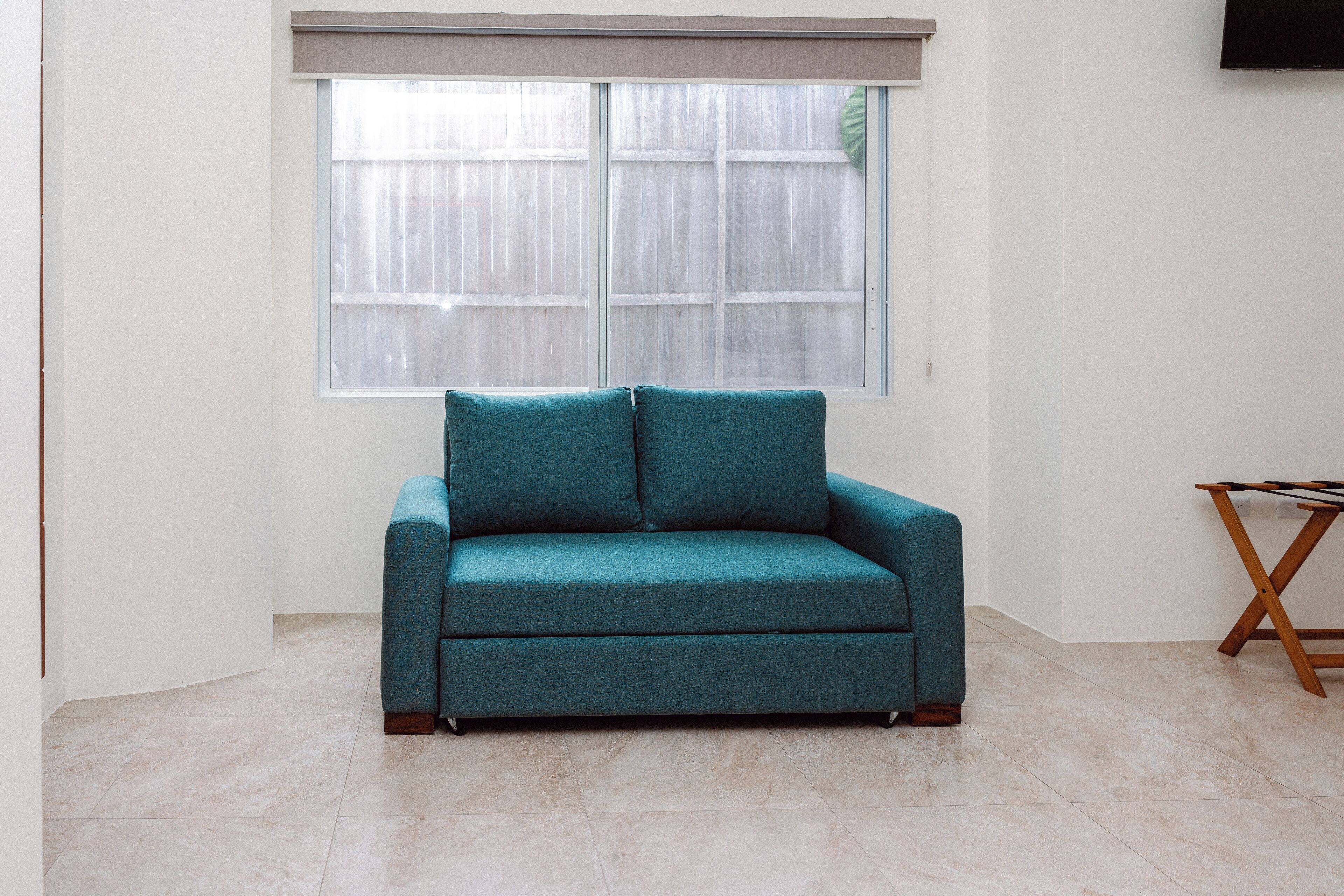 A teal sofa in a room with a large window, a wooden table, and a tiled floor.