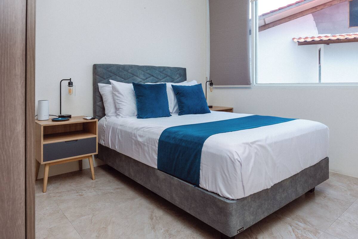 A neatly made bed with a grey headboard and blue pillows, a bedside table with a lamp, and a window with a view of a building.