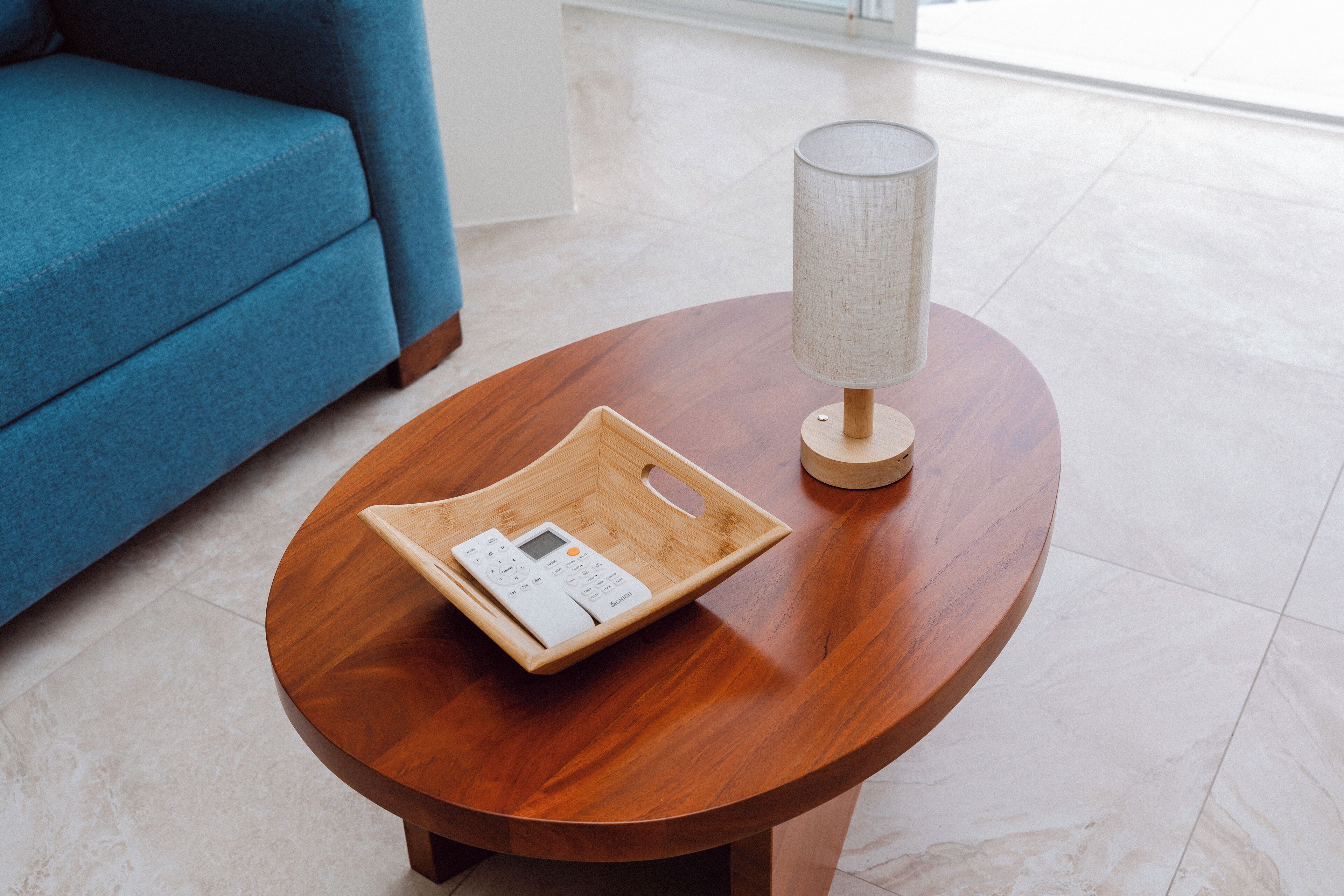 Una mesa redonda de madera con una bandeja que contiene un control remoto, una lámpara y un sofá azul al fondo.