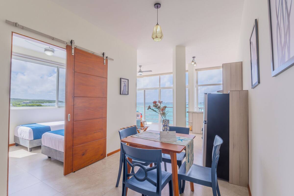 A room with a bed, a table with chairs, a view of the sea, and a sliding door.
