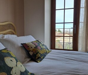A bedroom with a wicker headboard, a bed with white bedding, a colorful floral pillow, and a view through a large window.
