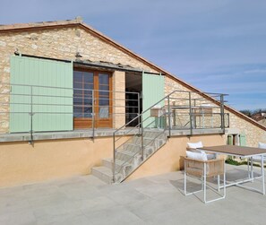 A stone building with a staircase leading to a balcony, a table with chairs, and a window with shutters.