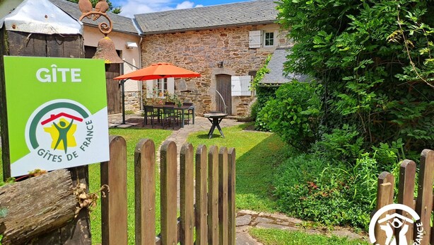 A stone building with a wooden fence, a patio with a table and chairs, and a sign reading "GÎTES DE FRANCE".