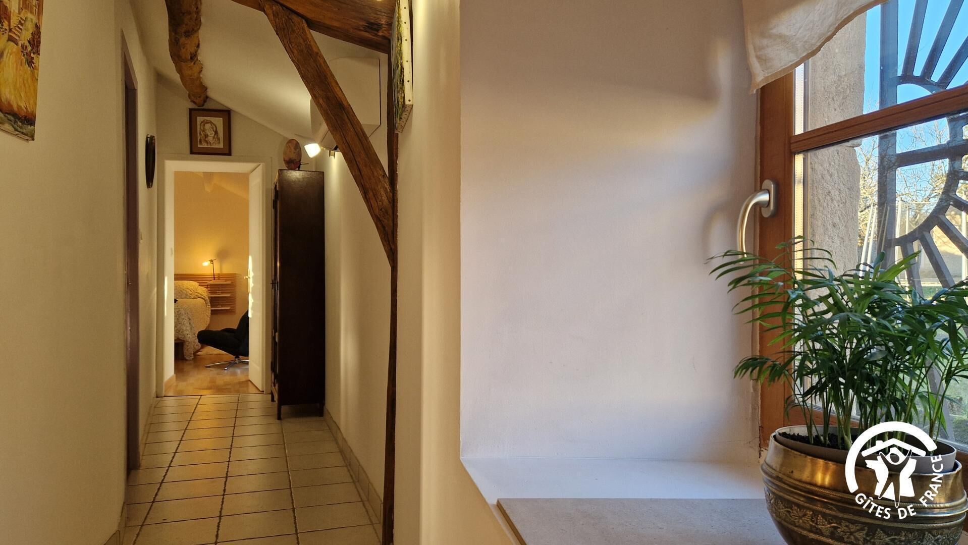 A narrow hallway with tiled flooring, wooden beams, and a potted plant on a windowsill.