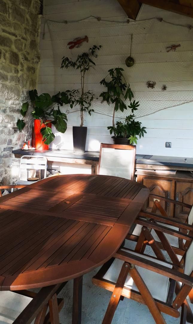 Une table à manger en bois, entourée de chaises, de plantes en pot et d’un mur de pierres.