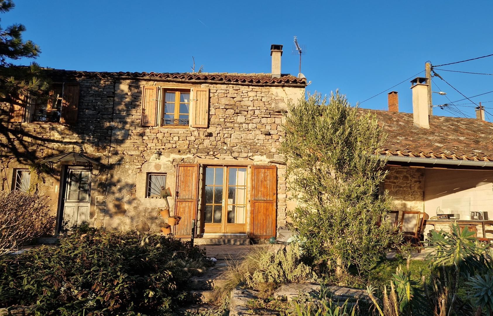 A stone house with a tiled roof, wooden shutters, and a chimney. There is a garden with various plants and a small table with items on it.