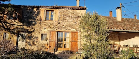 A stone house with a tiled roof, wooden shutters, and a chimney. There is a garden with various plants and a small table with items on it.