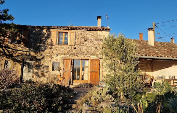 A stone house with a tiled roof, wooden shutters, and a chimney. There is a garden with various plants and a small table with items on it.