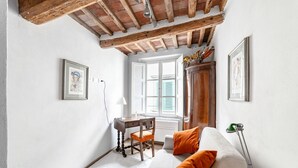 A room with a wooden ceiling, a white wall, a window with shutters, a desk with a chair, a sofa with orange cushions, and a lamp.