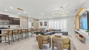 A modern kitchen with a central island, bar stools, a dining area with a sofa and chairs, and a living room with a large TV and a fireplace.