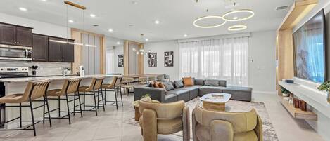 A modern kitchen with a central island, bar stools, a dining area with a sofa and chairs, and a living room with a large TV and a fireplace.
