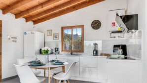 A modern kitchen with white cabinetry, a wooden ceiling, a window with a mountain view, and a dining area with white chairs and a table set for two. - Home2Book Country Stay with Terrace & BBQ (Las Palmas de Gran Canaria - Cuenca del Guiniguada)