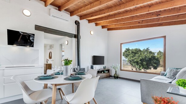 A modern kitchen with white cabinetry, a dining area with white chairs and a round table, a large window with a view of trees, and a grey sofa with a plant. - Home2Book Country Stay with Terrace & BBQ (Las Palmas de Gran Canaria - Cuenca del Guiniguada)