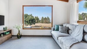 A living room with a large window, a grey sofa, a wooden TV stand, and a vase with flowers. - Home2Book Country Stay with Terrace & BBQ (Las Palmas de Gran Canaria - Cuenca del Guiniguada)