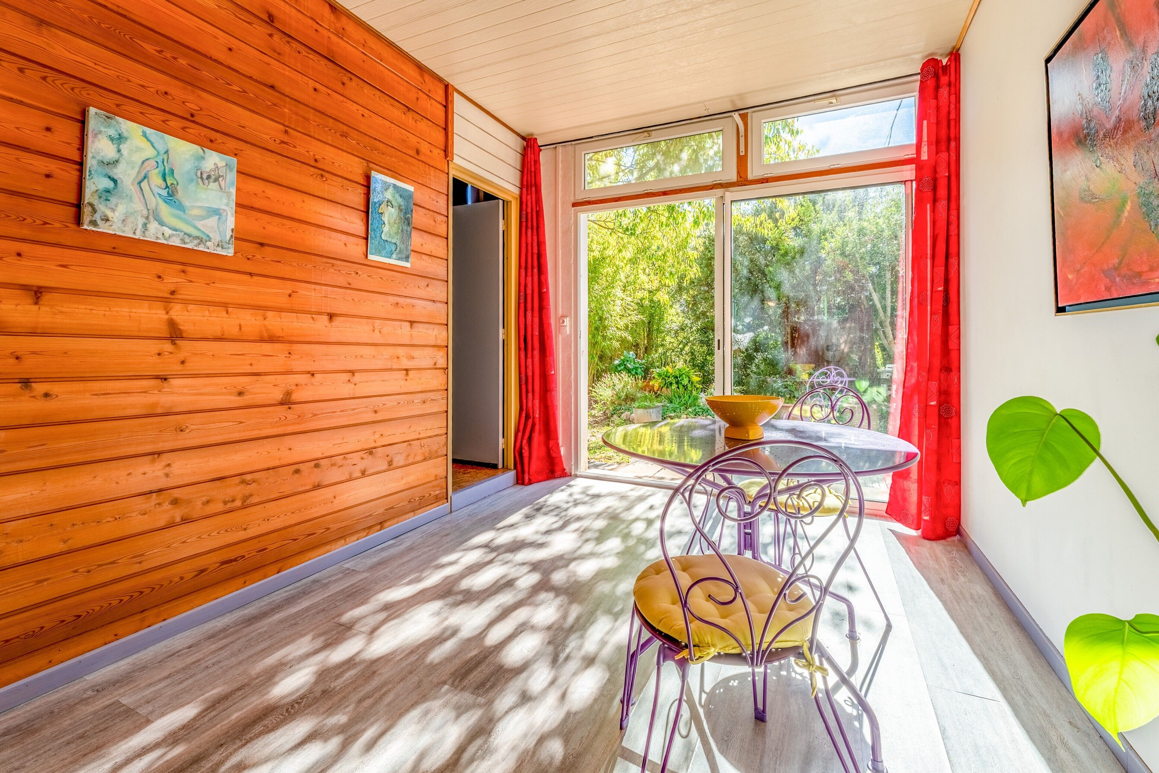 A room with wooden walls, a dining table with chairs, and a view of greenery outside.