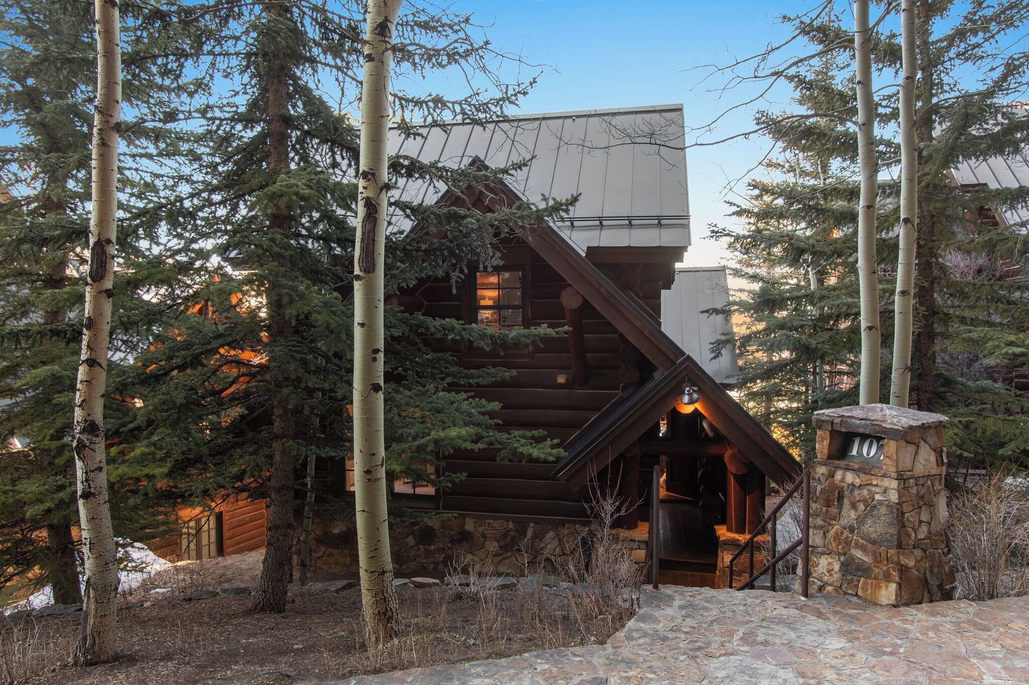 Une cabane en rondins avec un escalier en pierre menant à une porte en bois, entourée de grands arbres et d’un mur de pierre.