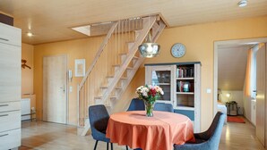 Een eetgedeelte met een tafel, stoelen, een vaas met bloemen, een trap en een boekenplank.