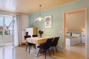 A dining area with a table, chairs, a hanging light fixture, a clock, and a bed in the adjacent room.