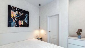Un dormitorio con una cama blanca, un cuadro en la pared, una mesita de noche con una lámpara y un aparador con un jarrón de flores.