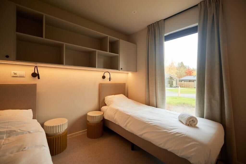 Ein Hotelzimmer mit zwei Betten, einem kleinen Hocker und einem Regal darüber.