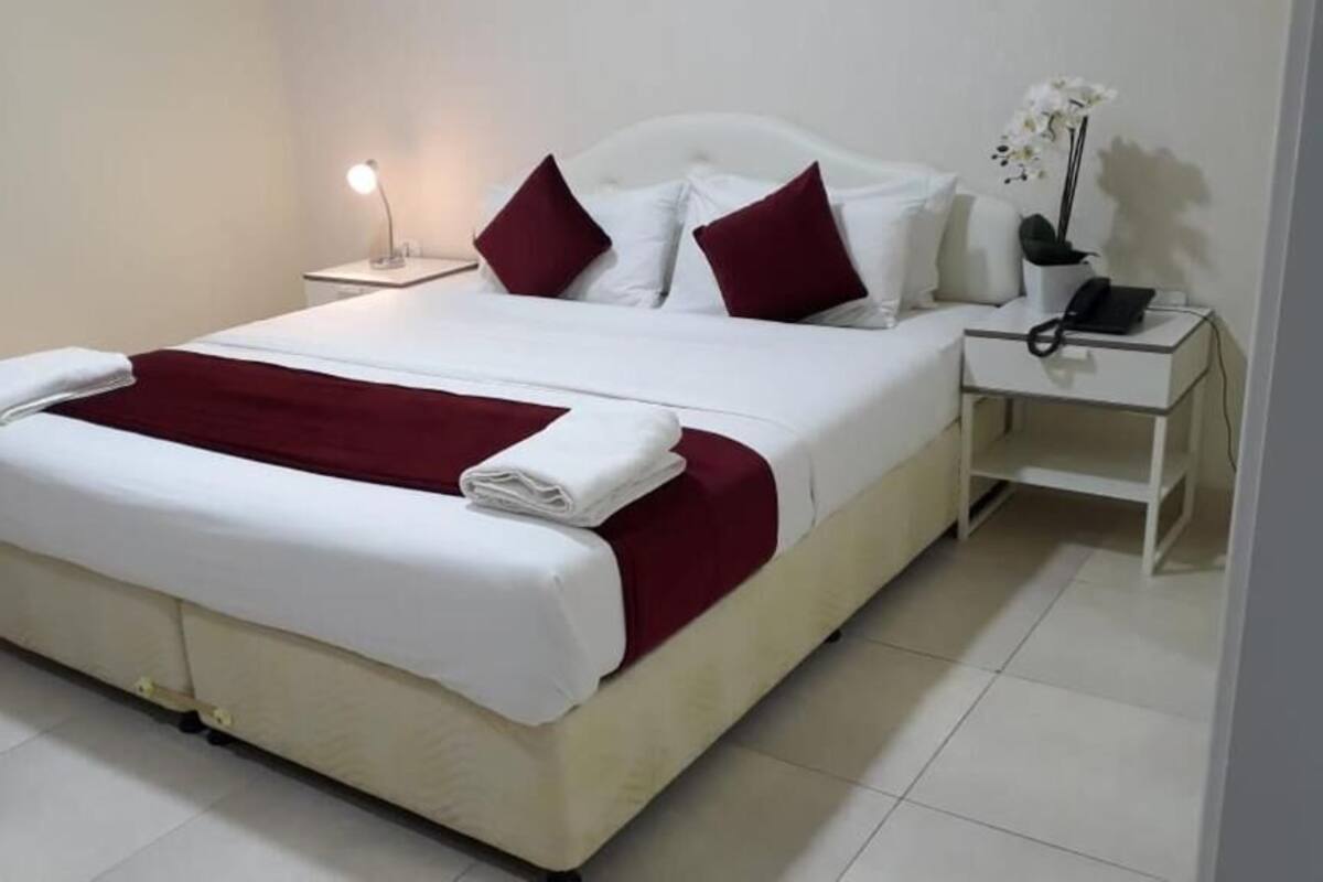 A neatly made bed with white and red bedding, a bedside lamp, a nightstand with a phone, and a small potted plant.