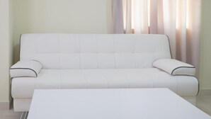 A white sofa with a matching ottoman in a room with light-colored walls and sheer curtains. - Al Jisr Hotel (Muscat)