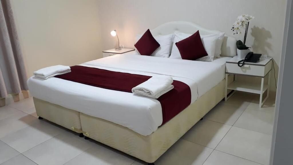 A neatly made bed with white and red bedding, a bedside lamp, a nightstand with a phone, and a small potted plant.