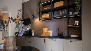 A modern kitchen with a dark countertop, white cabinets, and a black chair. There are wicker baskets on shelves, a vase with flowers, and a small plant. - Maison Amelie – Design Apartment | Milano Centrale (Milan)