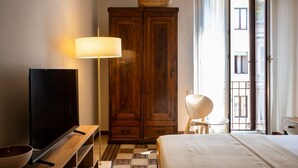 A hotel room with a television, a wooden wardrobe, a bedside lamp, a bed, and a view of the outside. - Maison Amelie – Design Apartment | Milano Centrale (Milan)