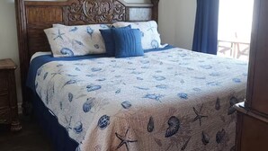 Un dormitorio con cabecera de madera, cama con colcha azul y blanca, y vista al exterior a través de una ventana con cortinas.