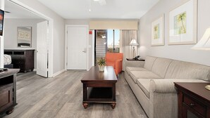 Un salón con una mesa de centro de madera oscura, un sofá beige y un sillón naranja. Se observa un ventilador de techo, una puerta que da a otra habitación y una escalera al fondo.