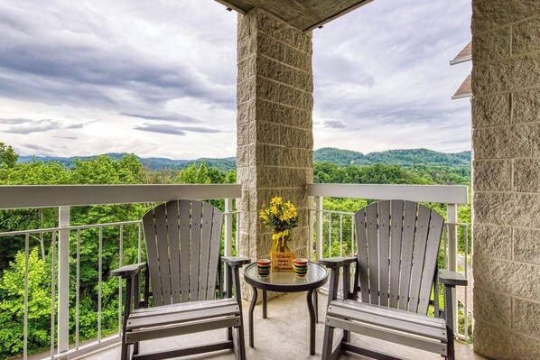 Ein Balkon mit zwei Adirondack-Stühlen, einem kleinen runden Tisch mit einer Vase voller gelber Blumen und einem Schild mit der Aufschrift 'Life is better in the Mountains'.