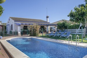 Piscina com espreguiçadeiras, alpendre coberto e edifício branco com telhado de azulejos.