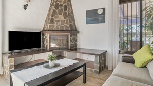 A living room with a stone fireplace, a flat-screen TV, a sofa, and a window with a view. - Holiday Home 'Isabel' with Private Pool, Wi-Fi and Air Conditioning (Conil de la Frontera)