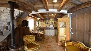 A rustic interior with exposed wooden beams, a stone fireplace, a dining area with a table and chairs, and a kitchen with wooden cabinets.