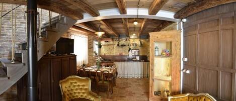 A rustic interior with exposed wooden beams, a stone fireplace, a dining area with a table and chairs, and a kitchen with wooden cabinets.