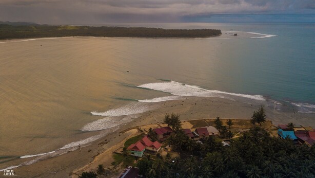 An aerial view of a coastal area with a beach, a small settlement with houses, and a forested landmass in the background.